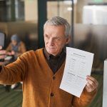 Homem idoso segura uma cédula e usa o celular para tirar uma selfie dentro de uma cabine de votação.
