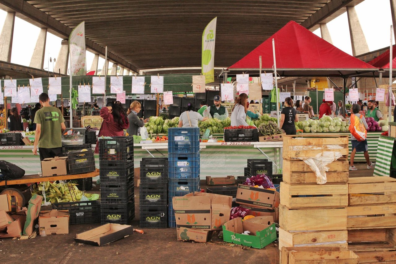 Pessoas fazem compras entre bancas de frutas e verduras em um mercado de São Paulo.