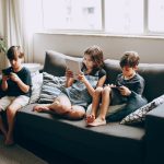 Três crianças sentadas em um sofá, concentradas no uso de dispositivos digitais dentro de casa.