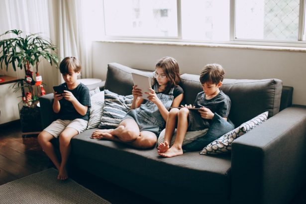 Três crianças sentadas em um sofá, concentradas no uso de dispositivos digitais dentro de casa.