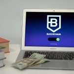 Laptop exibe uma interface de conexão blockchain sobre uma mesa com livros e dólares americanos.