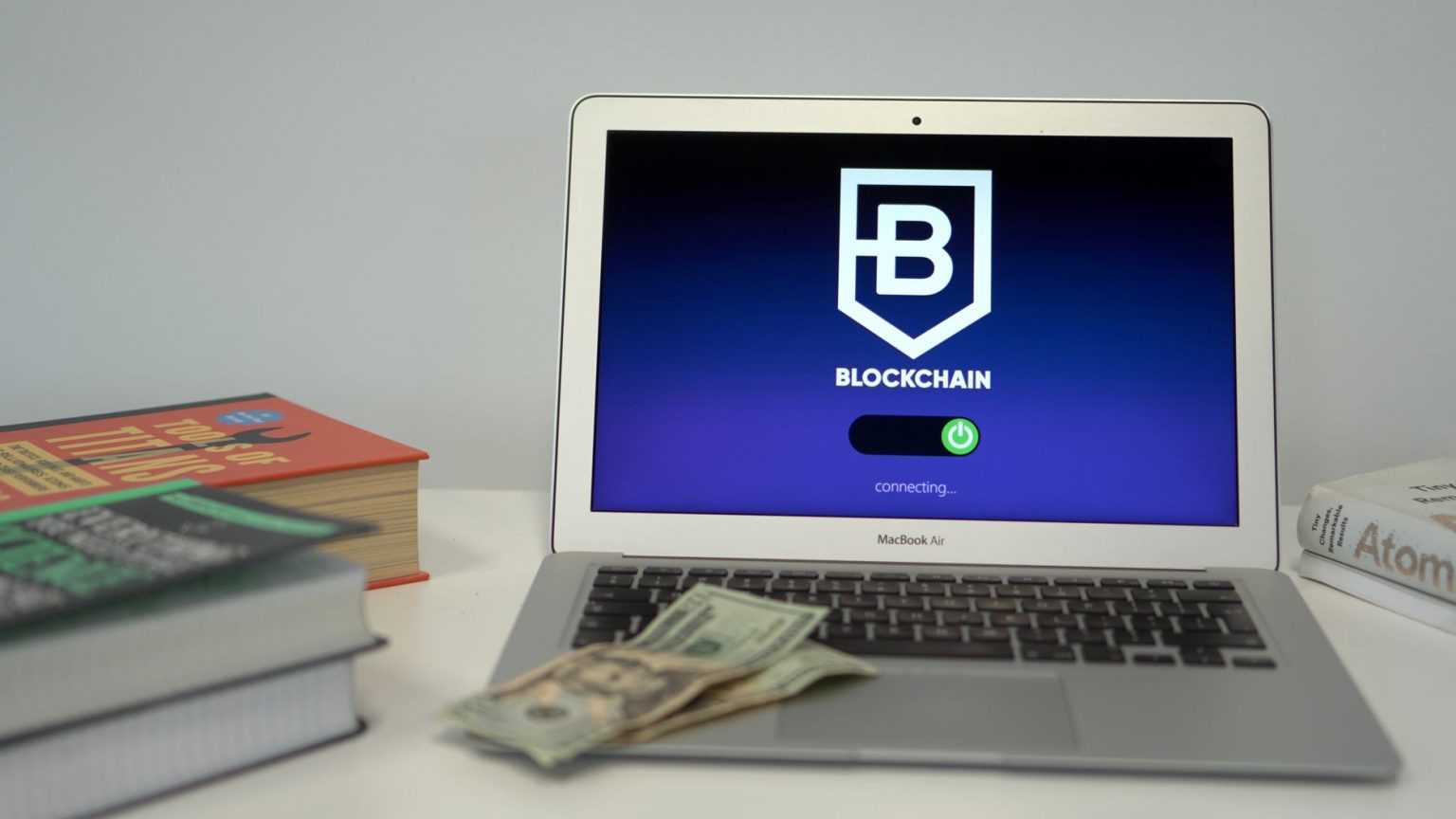 Laptop exibe uma interface de conexão blockchain sobre uma mesa com livros e dólares americanos.